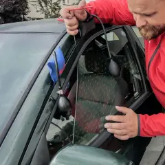 Mężczyzna w czerwonej kurtce używa narzędzi, aby otworzyć zatrzaśnięty samochód. Widać pompkę i cienki drut.