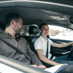 Ojciec z synem w samochodzie. Chłopak z zębami w aparacie prowadzi, a ojciec obok niego z uśmiechem. To może być ich pierwsza wspólna jazda po zdanym egzaminie teoretycznym na prawo jazdy.