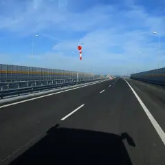 Pusta droga dwukierunkowa o dwóch pasach ruchu, otoczona barierami dźwiękochłonnymi. W oddali widać rękaw lotniczy.