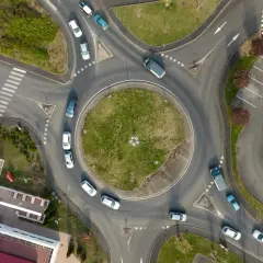 Widok z lotu ptaka na rondo, które jest skrzyżowaniem z ruchem okrężnym. Samochody poruszają się po okręgu, a obok znajduje się parking.
