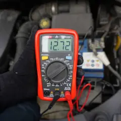 Ręka w czarnej rękawiczce trzyma czerwony miernik, który pokazuje 12.72V. To pokazuje, jak sprawdzić akumulator miernikiem.