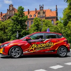 Czerwony samochód z napisem "Akademia dobrej jazdy" i niebieskim znakiem "L" na dachu. To idealny pojazd do nauki jazdy, który oferuje kompleksowy program szkoleniowy.
