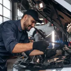 Uśmiechnięty mechanik samochodowy w granatowym kombinezonie sprawdza silnik. Zastanawiasz się, ile zarabia mechanik samochodowy?