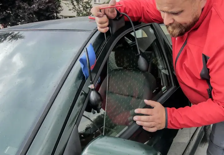 Mężczyzna w czerwonej kurtce używa narzędzi, aby otworzyć zatrzaśnięty samochód. Widać pompkę i cienki drut.