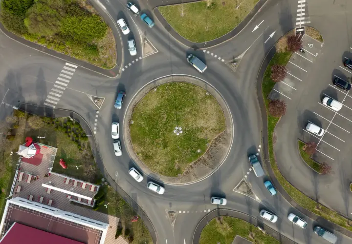 Widok z lotu ptaka na rondo, które jest skrzyżowaniem z ruchem okrężnym. Samochody poruszają się po okręgu, a obok znajduje się parking.