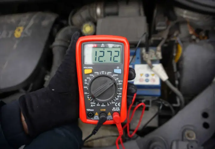 Ręka w czarnej rękawiczce trzyma czerwony miernik, który pokazuje 12.72V. To pokazuje, jak sprawdzić akumulator miernikiem.