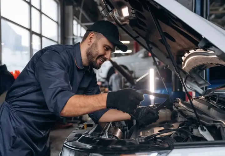 Uśmiechnięty mechanik samochodowy w granatowym kombinezonie sprawdza silnik. Zastanawiasz się, ile zarabia mechanik samochodowy?