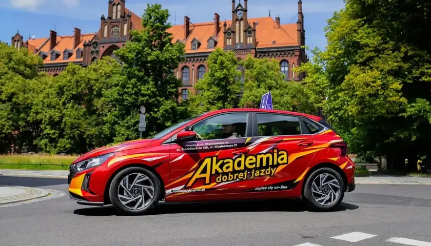 Czerwony samochód z napisem "Akademia dobrej jazdy" i niebieskim znakiem "L" na dachu. To idealny pojazd do nauki jazdy, który oferuje kompleksowy program szkoleniowy.
