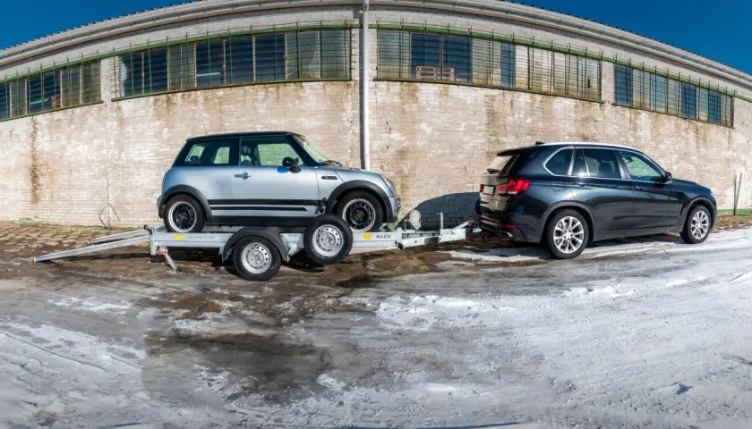 Mini Cooper na lawecie, obok BMW X5. Pokazano, jak zabezpieczyć auto na lawecie, używając pasów.