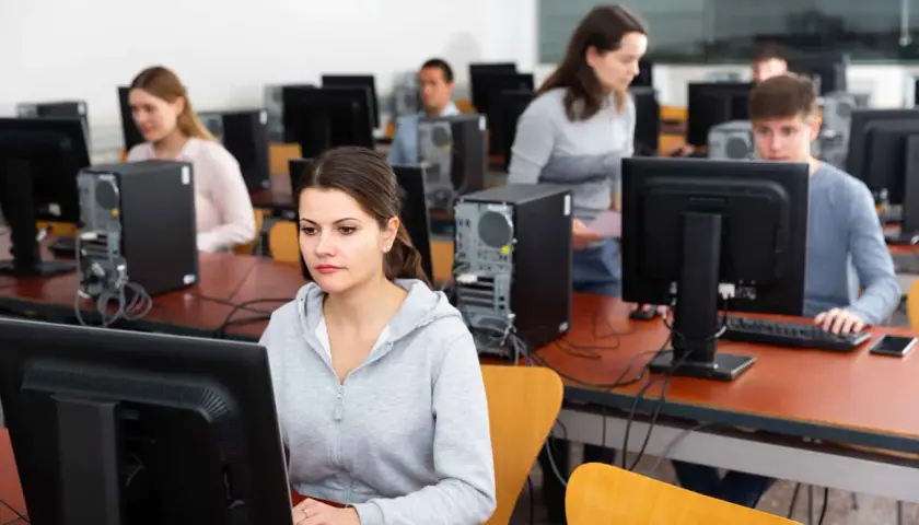 Uczniowie w sali komputerowej ćwiczą, jak zdać teorię na prawko. Skupieni przed monitorami, przygotowują się do egzaminu.