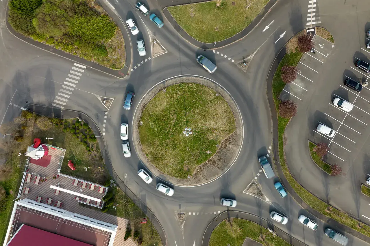 Widok z lotu ptaka na rondo, które jest skrzyżowaniem z ruchem okrężnym. Samochody poruszają się po okręgu, a obok znajduje się parking.