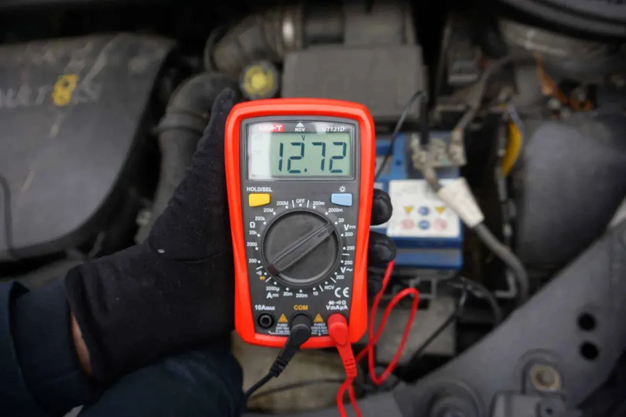 Ręka w czarnej rękawiczce trzyma czerwony miernik, który pokazuje 12.72V. To pokazuje, jak sprawdzić akumulator miernikiem.