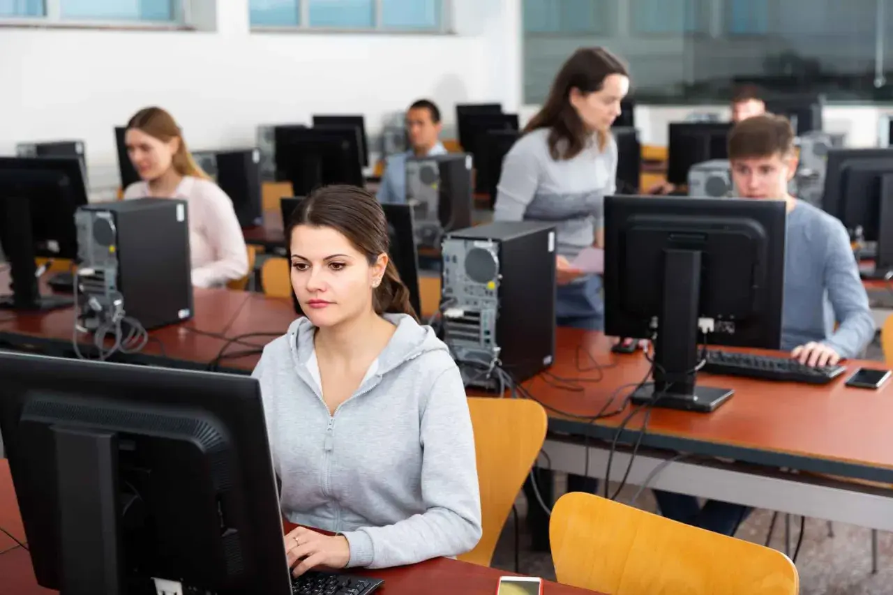 Uczniowie w sali komputerowej ćwiczą, jak zdać teorię na prawko. Skupieni przed monitorami, przygotowują się do egzaminu.