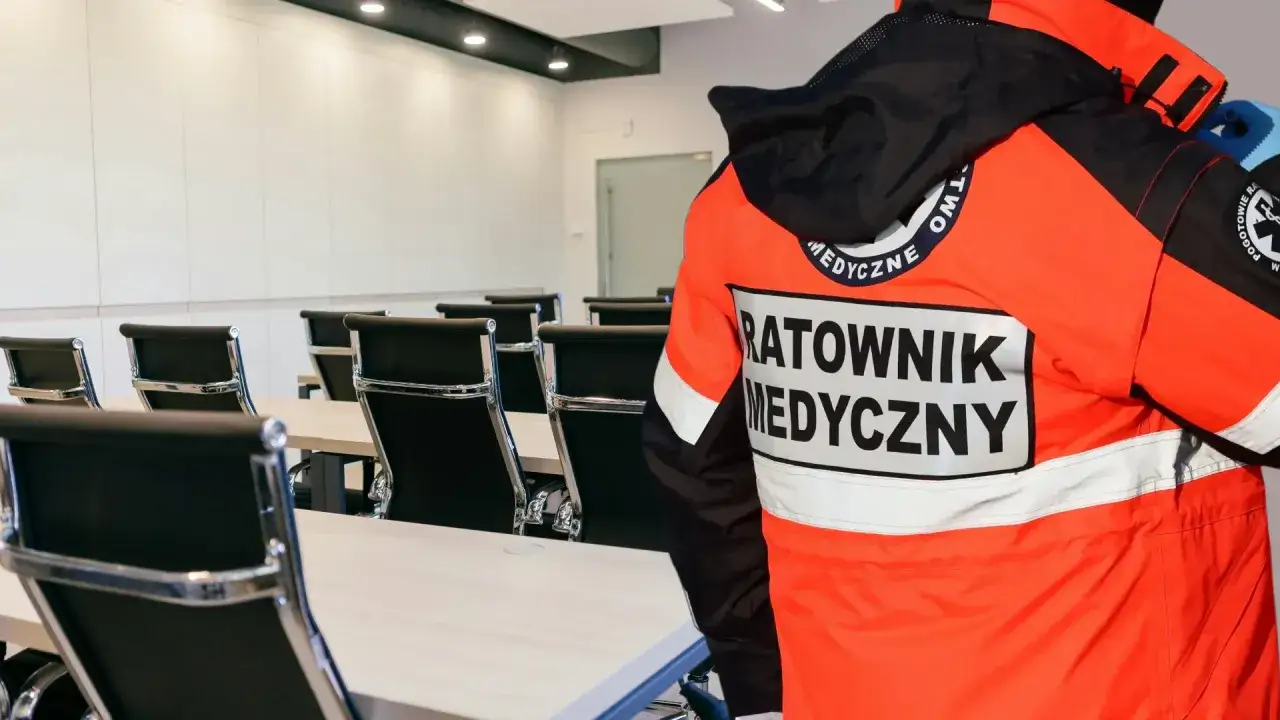 Ratownik medyczny w pomarańczowej kurtce stoi w sali konferencyjnej. Zastanawiasz się, ile trwa kurs pierwszej pomocy na prawo jazdy?