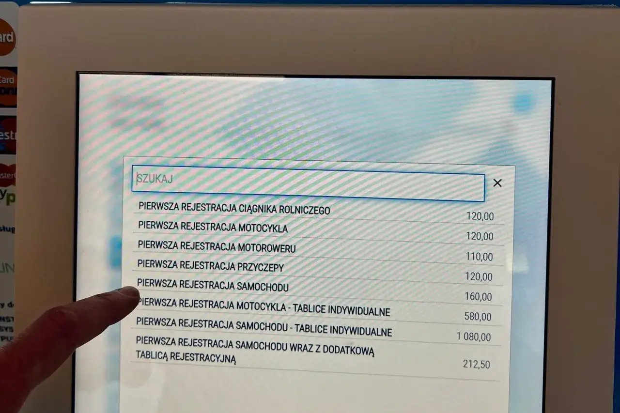 Palec wskazuje na ekran z listą usług, gdzie szukamy informacji, ile trwa przerejestrowanie auta. Widoczne są ceny rejestracji różnych pojazdów.