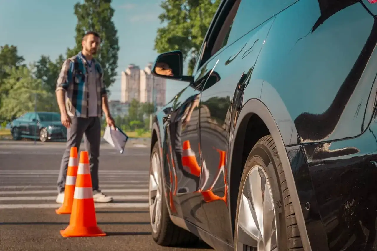 Egzaminator z dokumentami stoi obok czarnego samochodu na placu manewrowym. Czy można zobaczyć nagranie z egzaminu na prawo jazdy?