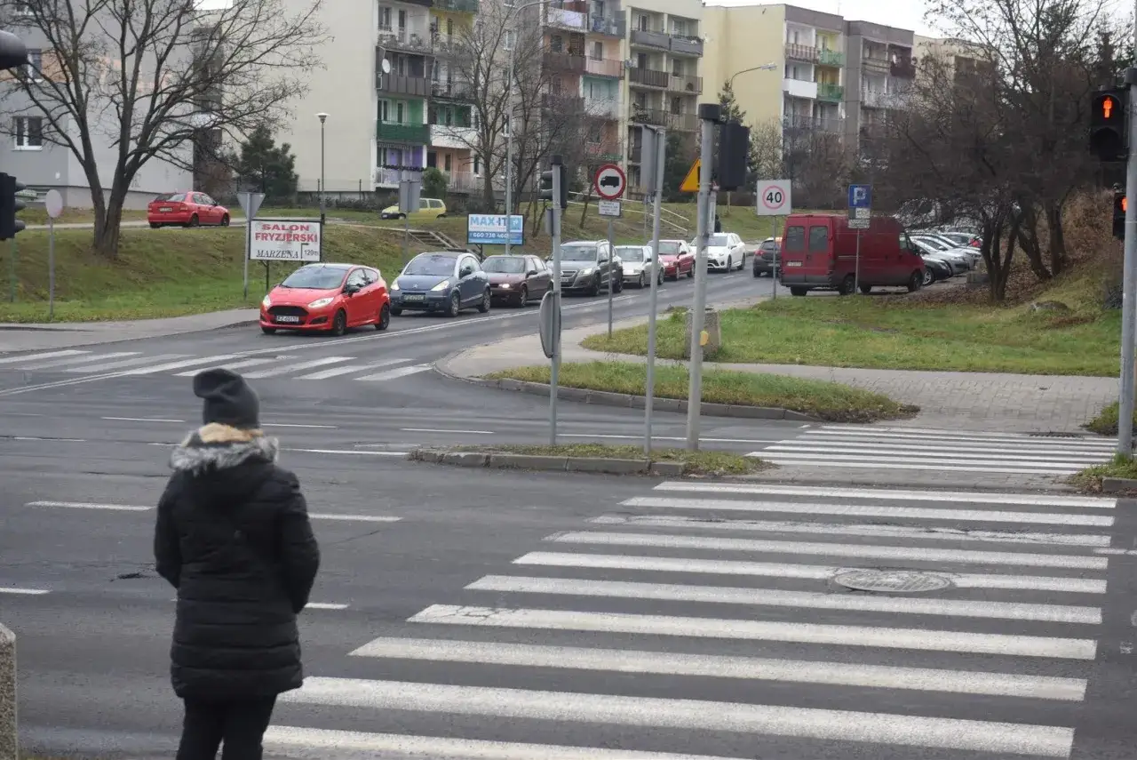 Kobieta czeka na zielone światło przy pasach, zgodnie z kodeksem drogowym. Samochody stoją w korku.