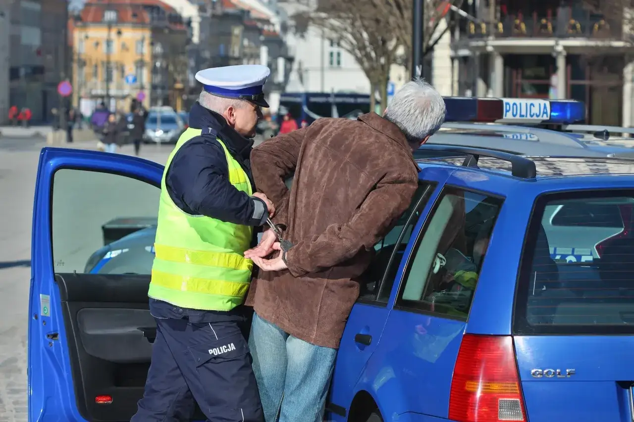 Policjant zakłada kajdanki mężczyźnie przy radiowozie. Może to być kara za jazdę bez prawa jazdy be egzamin.
